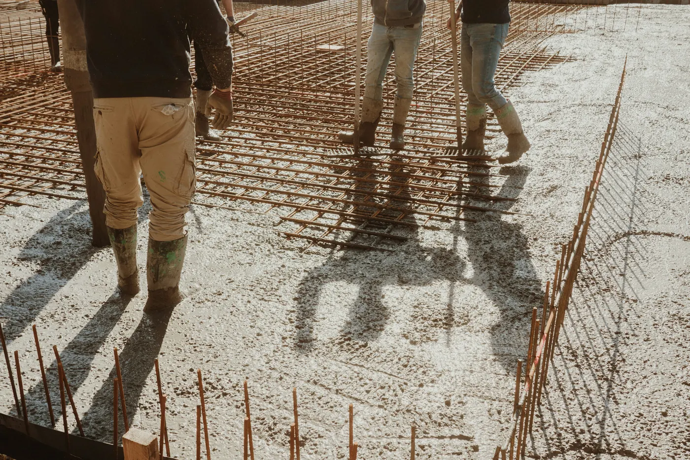 Workers in rubber boots spreading wet concrete across steel mesh for a new slab
