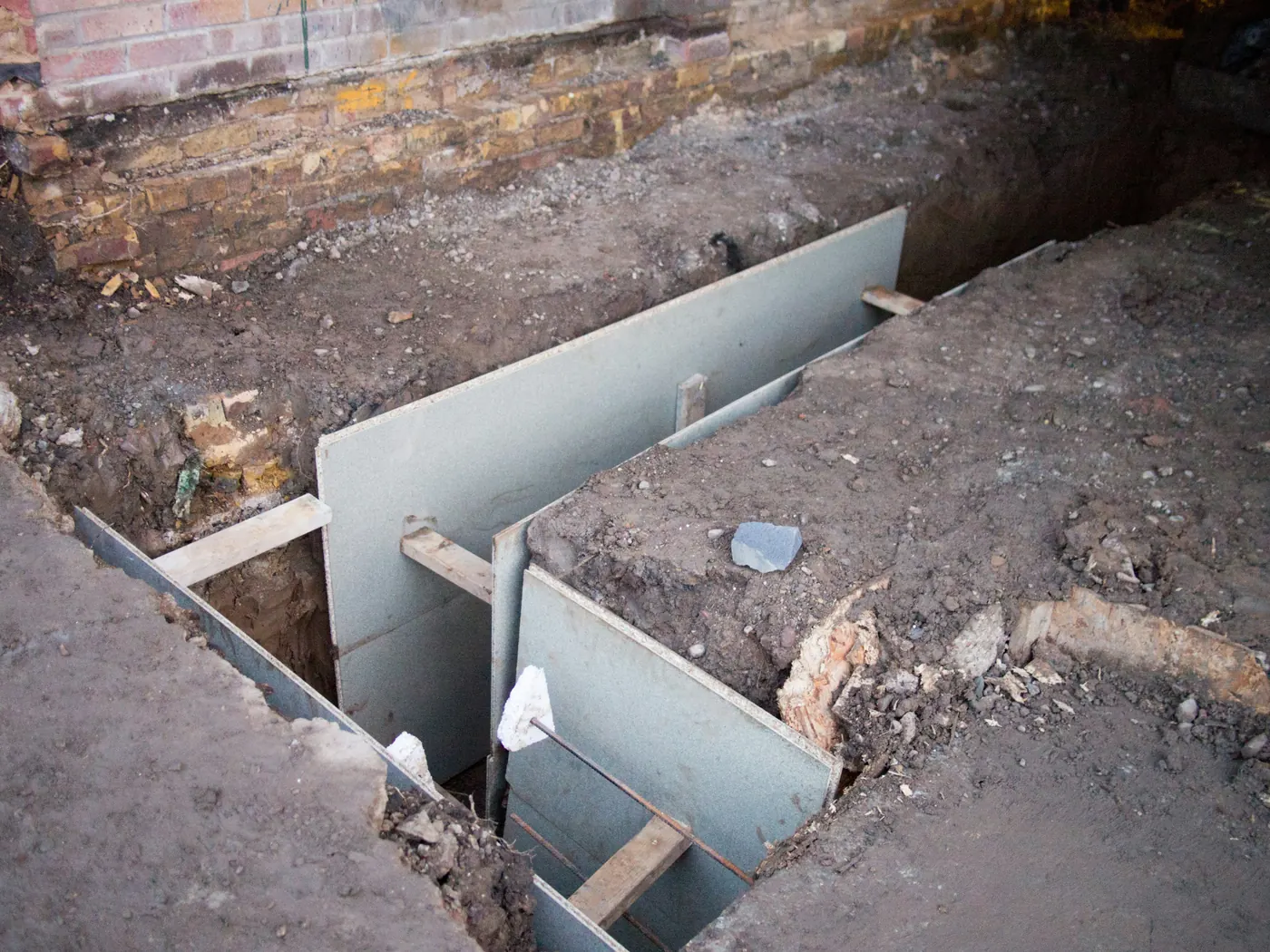 Narrow trench dug along the base of an old brick wall, lined with boards and timber braces